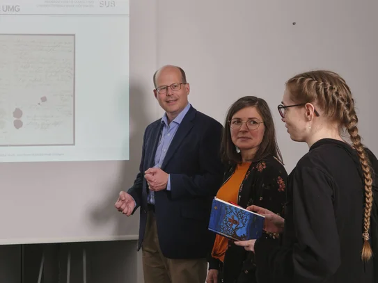 Vor einer Powerpointpräsentation stehen auf jeder Seite drei Menschen, die die Powerpoint betrachten. Diese zeigt eine historische Urkunde.