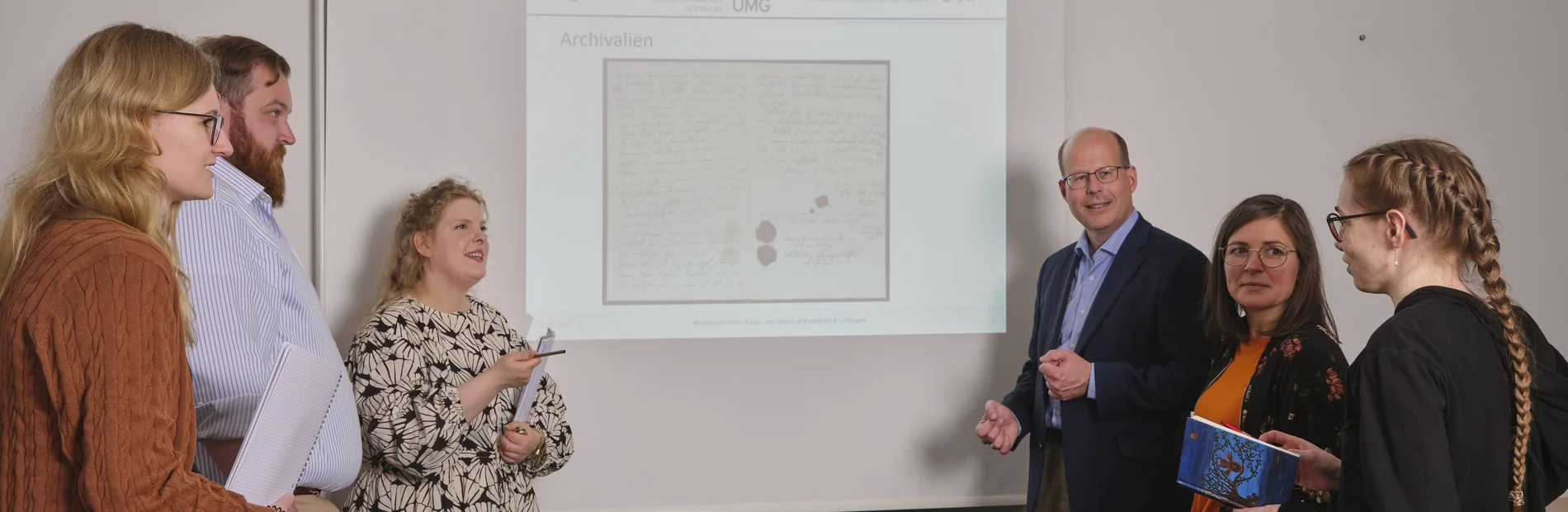 Kurse zur Arbeit mit historischen Beständen Vor einer Powerpointpräsentation stehen auf jeder Seite drei Menschen, die die Powerpoint betrachten. Diese zeigt eine historische Urkunde.