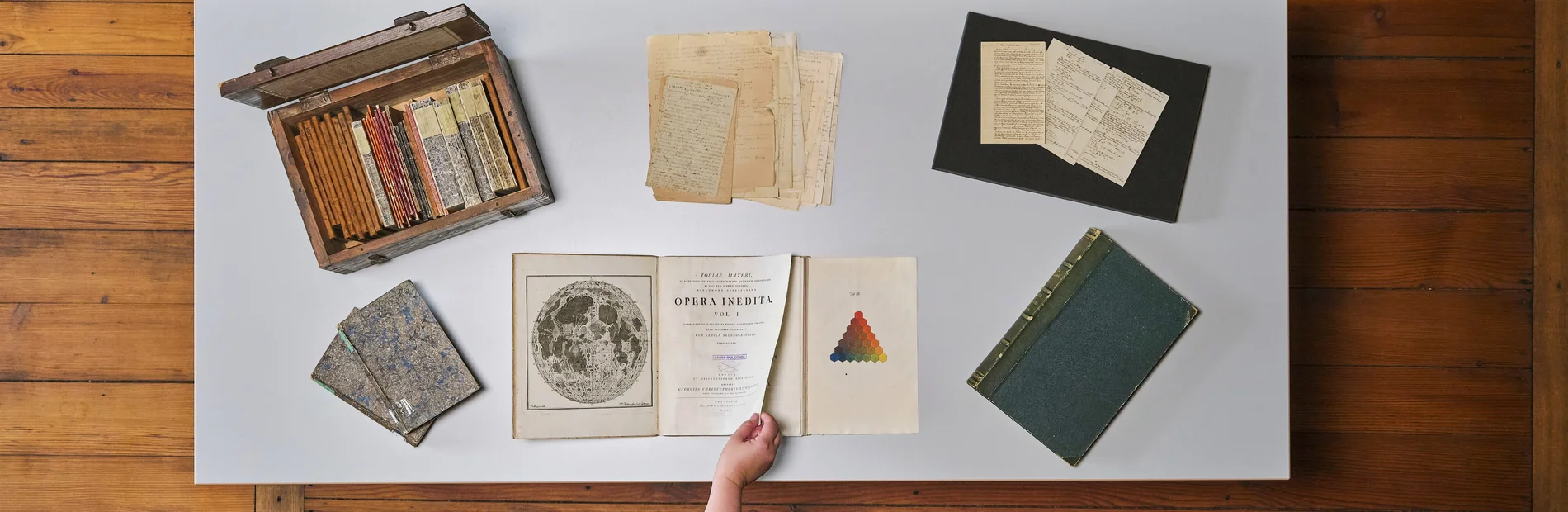 Tisch mit historischen Büchern und Archivmaterialien, eine Hand blättert in einem Buch