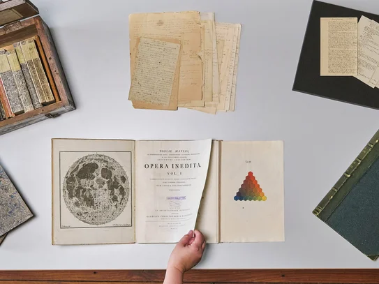 Tisch mit historischen Büchern und Archivmaterialien, eine Hand blättert in einem Buch
