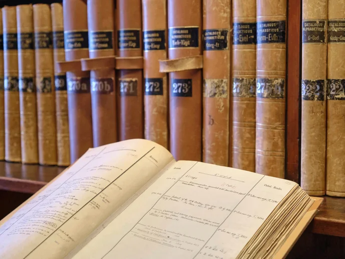 Im Hintergrund Bibliothekskataloge auf einem Regal, im linken vorderen Bereich ein aufgeschlagener Bibliothekskatalog