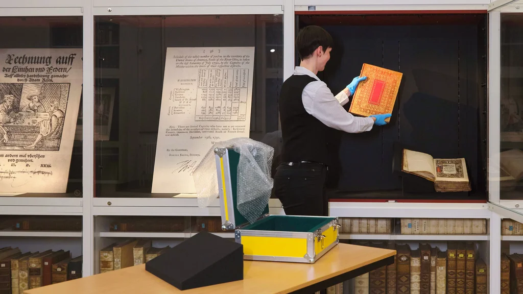 Restauratorin legt ein Buch in eine offene Vitrine; daneben geschlossene Vitrinen mit Faksimiles.