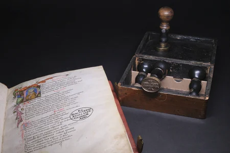 Historische Bibliotheksstempel Links eine aufgeschlagene Handschrift mit dem Stempel „EX BIBLIOTHECA REGIA ACAD. REGIAE AUG:“, rechts ein Holzkasten mit historischen Stempelwerkzeugen.
