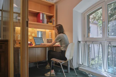 Das Bild zeigt das Innere einer Arbeitskabine. Eine Studentin sitzt an einem Tisch und benutzt einen Laptop. Auf dem Tisch sind verschiedene Arbeitsmaterialien verteilt. Auf dem Regal über dem Tisch stehen mehrere Ordner und Bücher.