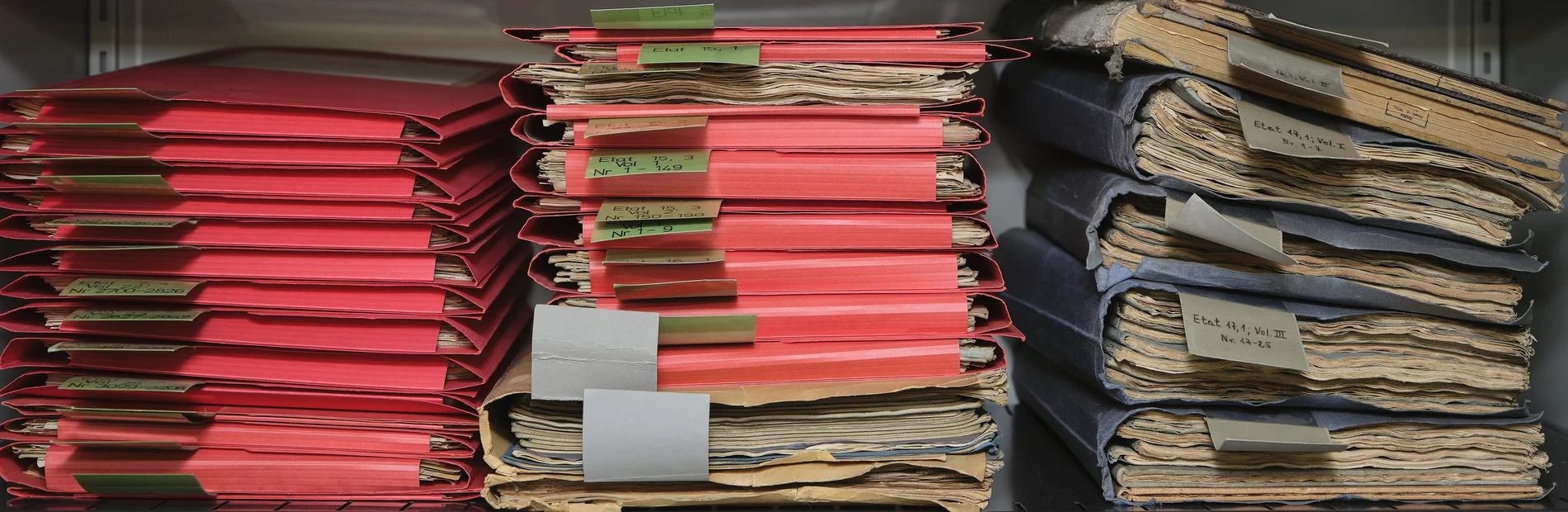 Drei Stapel mit roten und blauen Mappen, die historische Materialien enthalten