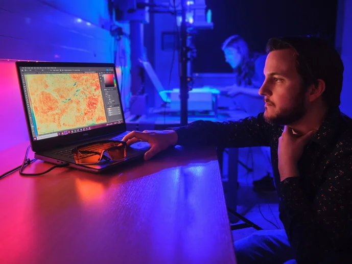 In pink- und violettfarbenem Licht bedient ein Mitarbeiter einen Computer, während im Hintergrund eine Mitarbeiterin eine Handschrift auf einer MSI-Anlage positioniert.