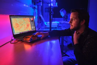 Digitale Rekonstruktion und Materialanalytik In pink- und violettfarbenem Licht bedient ein Mitarbeiter einen Computer, während im Hintergrund eine Mitarbeiterin eine Handschrift auf einer MSI-Anlage positioniert.