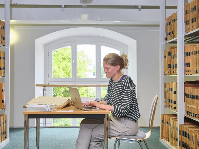 Nutzerin sitzt an einem Tisch zwischen Bücherregalen und tippt auf einem Laptop.