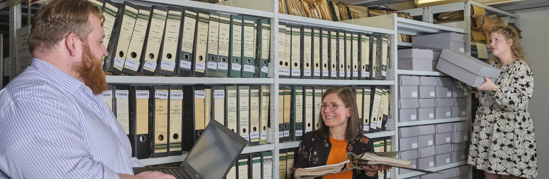 Drei Personen vor einem Regal mit Archivmaterial, eine Person nimmt eine Box heraus.