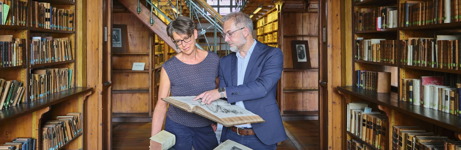 Zwei Personen im historischen Bibliothekssaal, eine zeigt der anderen ein altes Buch