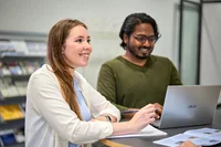 Zwei Personen stehen an einem Tisch, darauf steht ein Laptop. Eine Person hört aufmerksam zu